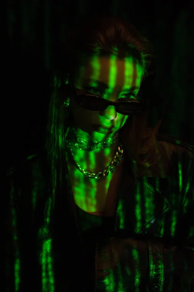 Young woman in dark sunglasses and metal accessories in green neon light isolated on black — стоковое фото