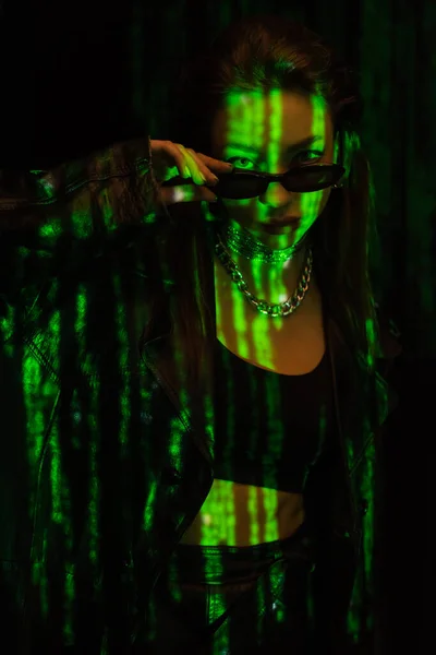 Young woman in silver necklaces holding dark sunglasses and looking at camera in green neon light isolated on black — Photo de stock