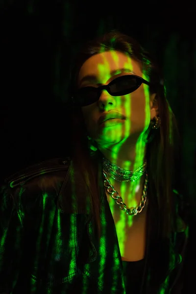 Portrait of trendy woman in dark sunglasses and silver necklaces in green neon light isolated on black — Photo de stock