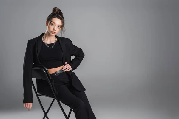 Young brunette woman in black trendy suit sitting on chair with hand on hip on grey background — Foto stock