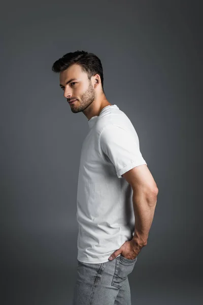Brunette man in white t-shirt holding hand in pocket of jeans isolated on grey — Stockfoto