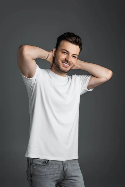 Positive man in white t-shirt looking at camera while touching neck isolated on grey — Stockfoto