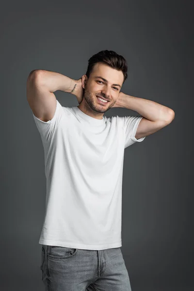 Positive man in white t-shirt looking at camera while touching neck isolated on grey — Foto stock