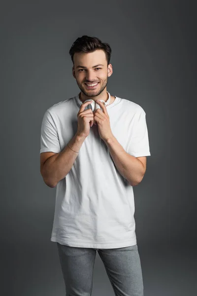 Positive man holding headphones and looking at camera isolated on grey — Foto stock