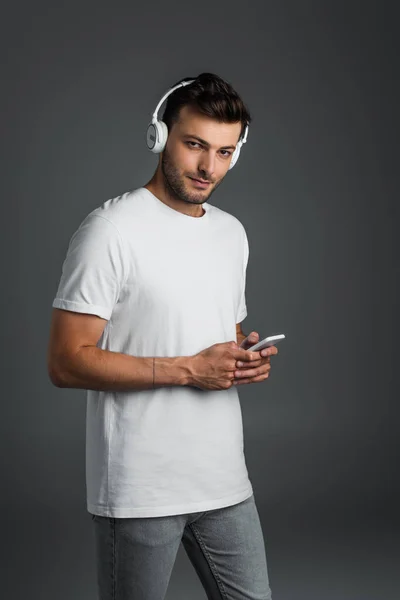 Молодий чоловік використовує бездротові навушники і смартфон і дивиться на камеру ізольовано на сірому — стокове фото