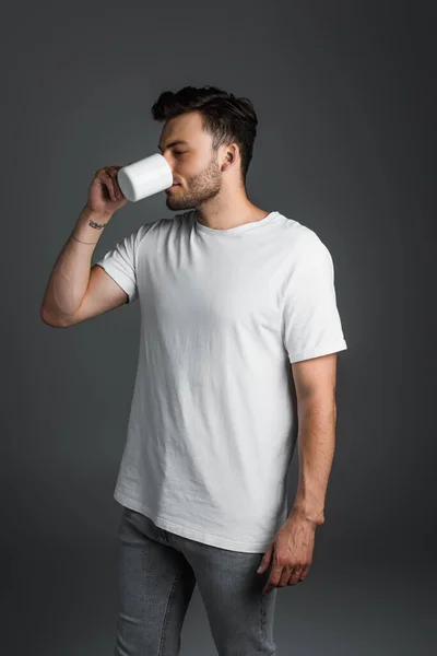 Молодий чоловік в джинсах і футболці п'є каву ізольовано на сірому — стокове фото