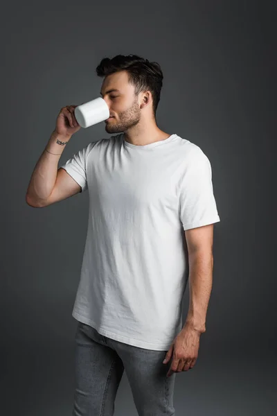 Young man in jeans and t-shirt drinking coffee isolated on grey — Fotografia de Stock