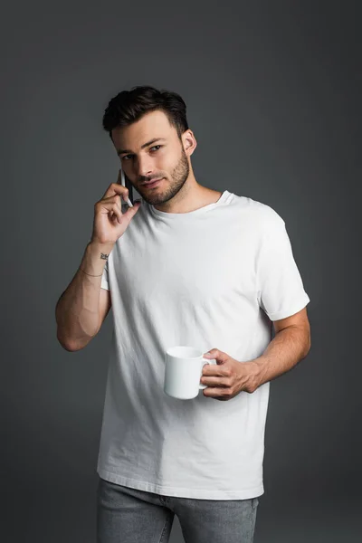 Brunette man holding cup of coffee while talking on smartphone isolated on grey — Stockfoto