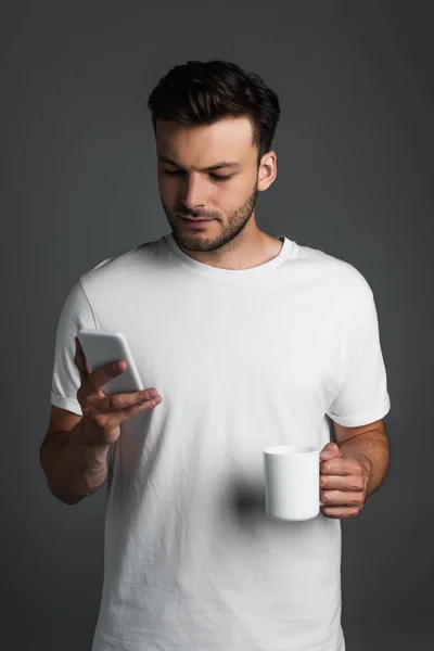 Молодой человек держит чашку кофе и с помощью смартфона изолированы на серый — стоковое фото