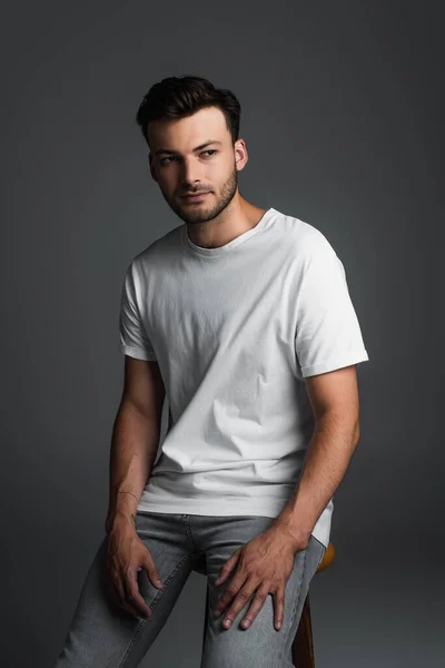 Bearded man in jeans and t-shirt sitting on chair isolated on grey — Stockfoto