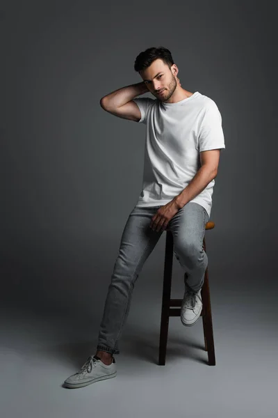 Full length of young man in t-shirt and jeans sitting on chair on grey background — Stock Photo