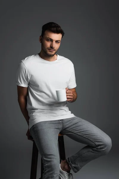 Молодий чоловік у білій футболці та джинсах тримає чашку кави, сидячи на стільці ізольовано на сірому — стокове фото