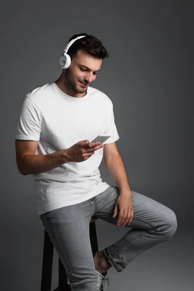 Smiling man using headphones and smartphone while sitting on chair isolated on grey — Stockfoto