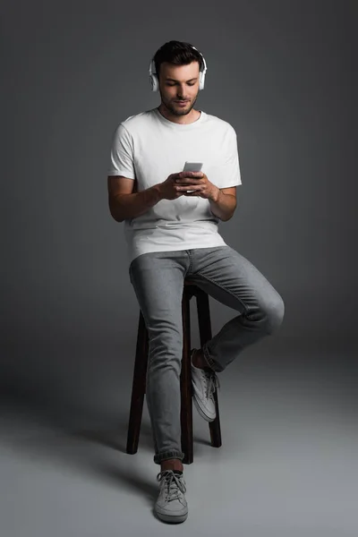 Full length of young man in jeans and t-shirt using smartphone and headphones while sitting on chair on grey background — Foto stock