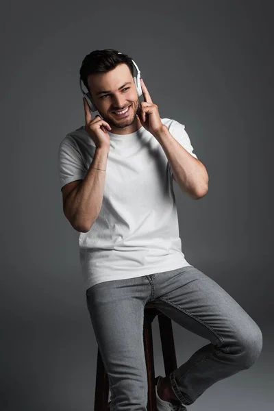 Smiling man in wireless headphones sitting on chair isolated on grey — Foto stock