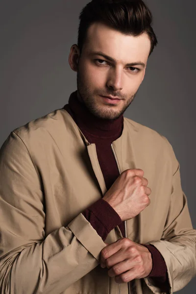 Portrait of trendy man adjusting autumn jacket and looking at camera isolated on grey — Fotografia de Stock