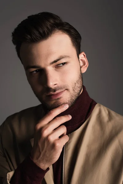 Brunette man in jacket touching chin and looking away isolated on grey - foto de stock