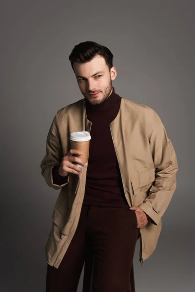 Стильний чоловік в коричневій куртці тримає каву в паперовій чашці ізольовано на сірому — стокове фото