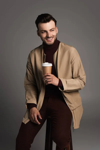 Smiling man in autumn outfit holding coffee to go while sitting on chair isolated on grey — Photo de stock