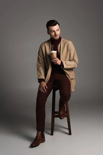 Повна довжина стильного чоловіка в осінньому вбранні тримає паперову чашку, сидячи на стільці на сірому фоні — стокове фото