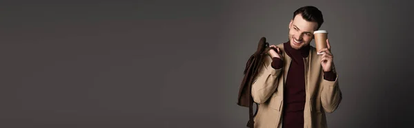 Веселый мужчина в осенней куртке держит рюкзак и кофе, чтобы пойти изолированы на серый, баннер — стоковое фото