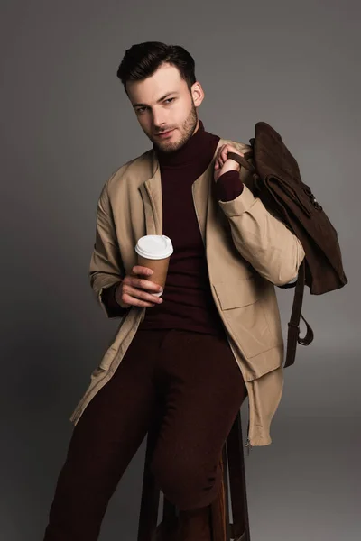 Stylish model in autumn outfit holding paper cup and backpack while sitting on chair isolated on grey — Stock Photo