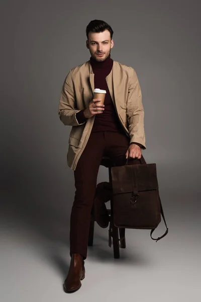 Trendy man in autumn jacket holding backpack and coffee to go while posing on chair on grey background — Photo de stock