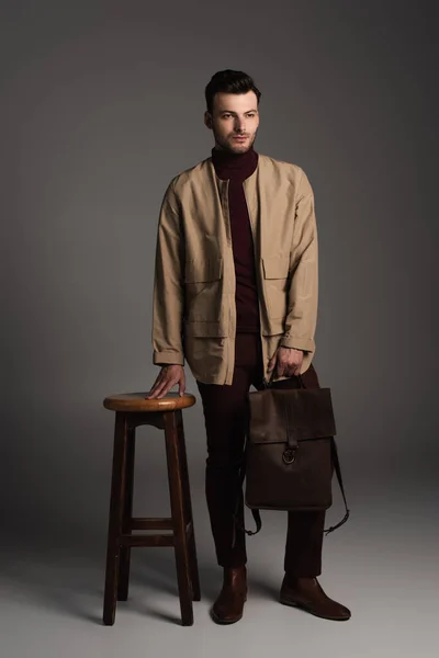 Full length of bearded man in autumn outfit holding backpack near chair on grey background - foto de stock