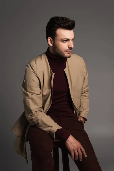 Trendy brunette man in brown jacket sitting on chair isolated on grey — Photo de stock