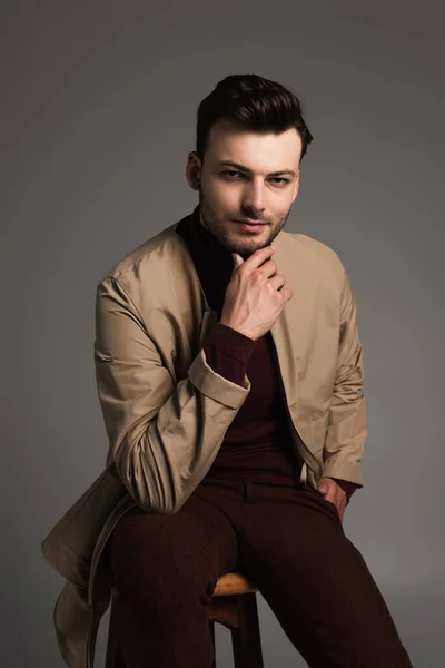 Portrait of stylish man in turtleneck and jacket sitting on chair and posing isolated on grey — Stockfoto