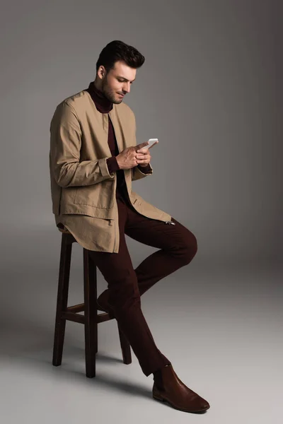 Young man in autumn outfit using smartphone while sitting on chair on grey background — Stock Photo