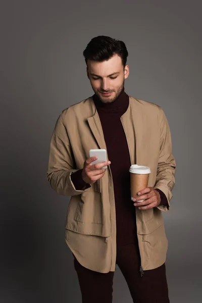Стильный мужчина в коричневой куртке, используя мобильный телефон и держа на вынос напиток, изолированный на сером — стоковое фото