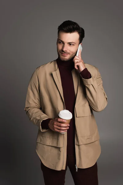 Бородатый мужчина в осеннем наряде разговаривает по мобильному телефону и держит кофе, чтобы пойти изолированы на серый — стоковое фото