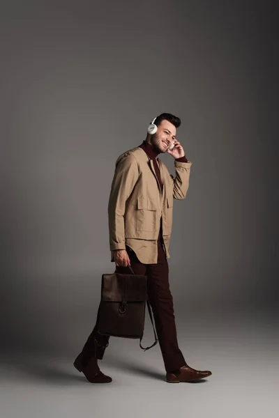 Smiling man in autumn outfit using headphones and holding backpack on grey background - foto de stock