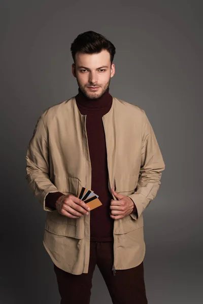 Brunette man in autumn outfit holding credit cards and looking at camera isolated on grey — Stockfoto