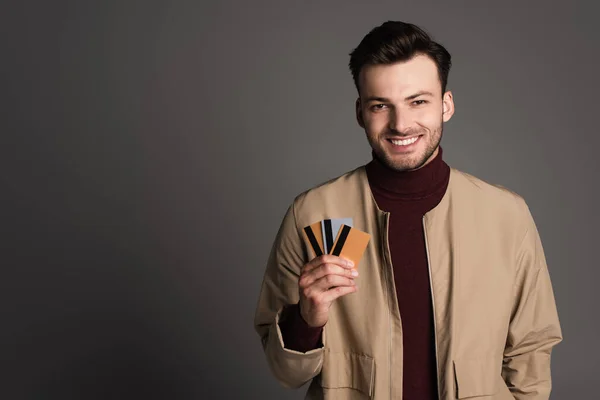 Cheerful man in autumn jacket holding credit cards isolated on grey — Fotografia de Stock