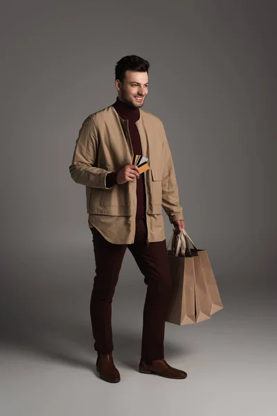 Full length of smiling man in autumn outfit holding shopping bags and credit cards on grey background - foto de stock