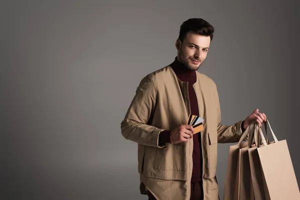 Чоловік в коричневій куртці тримає сумки і кредитні картки ізольовані на сірому — стокове фото