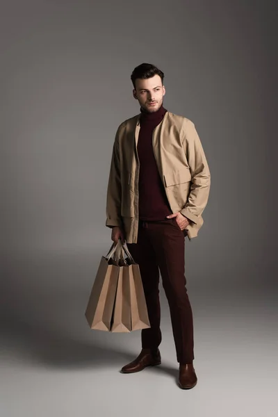 Full length of stylish man in autumn outfit holding shopping bags on grey background — Stock Photo