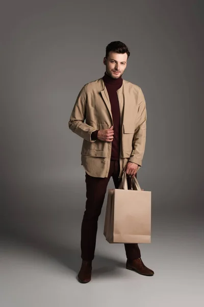 Fashionable man in brown jacket holding shopping bags on grey background — Stockfoto