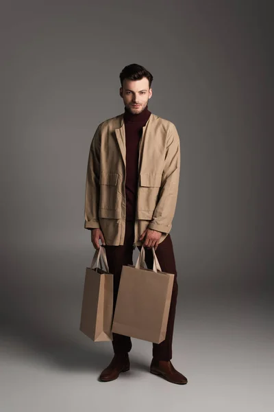 Full length of brunette man in autumn outfit holding purchases on grey background — Photo de stock