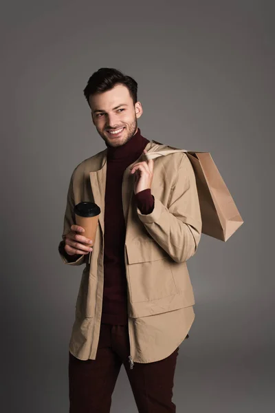 Smiling man in brown jacket holding coffee in paper cup and shopping bag isolated on grey — Foto stock