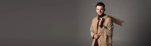 Smiling man in autumn jacket holding purchase and looking at camera isolated on grey, banner — Fotografia de Stock