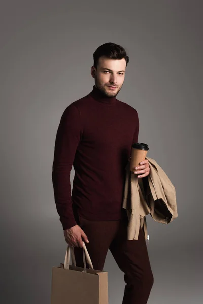 Стильний брюнетка чоловік з піджаком тримає каву, щоб піти і торгувати сумкою ізольовано на сірому — стокове фото