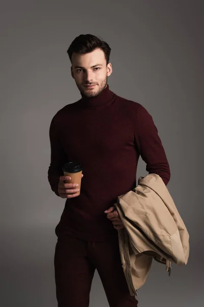 Trendy young man holding autumn jacket and paper cup isolated on grey — Stockfoto