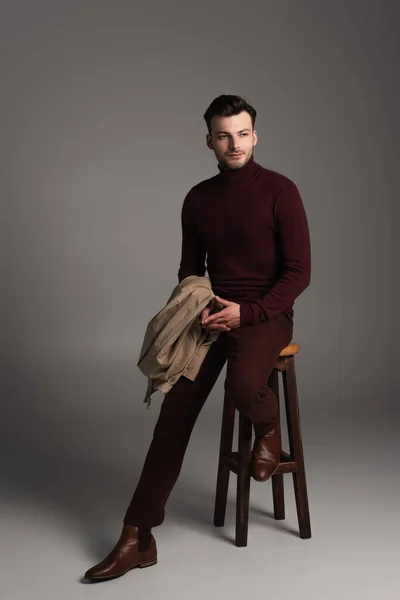 Full length of young barded man in turtleneck holding jacket while sitting on chair on grey background - foto de stock