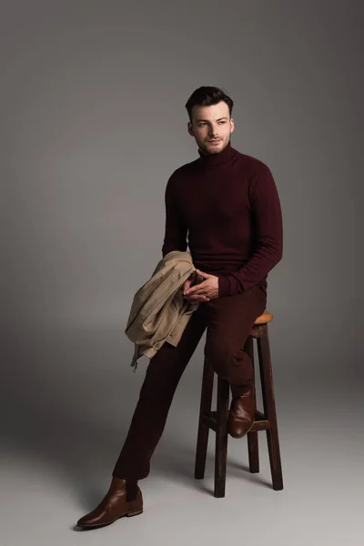 Full length of young barded man in turtleneck holding jacket while sitting on chair on grey background — Stock Photo