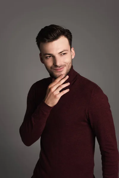 Smiling brunette man in turtleneck touching chin isolated on grey — Stockfoto