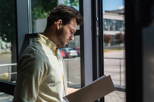 Вид сбоку на бизнесмена, смотрящего на бумажную папку у окна офиса — стоковое фото