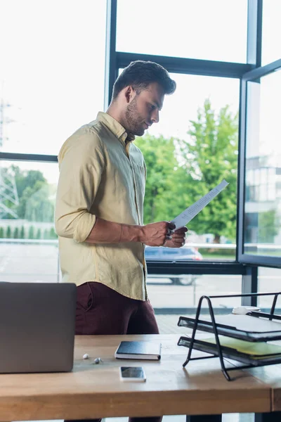 Side view of businessman in shirt holding document near devices and papers in office - foto de stock